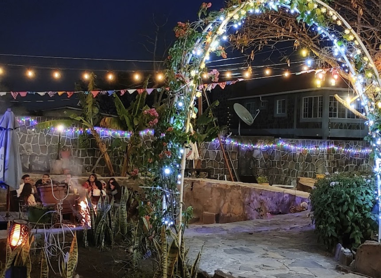 Arco decorativo con luces de hada, plantas y visitantes reunidos junto a la fogata.
