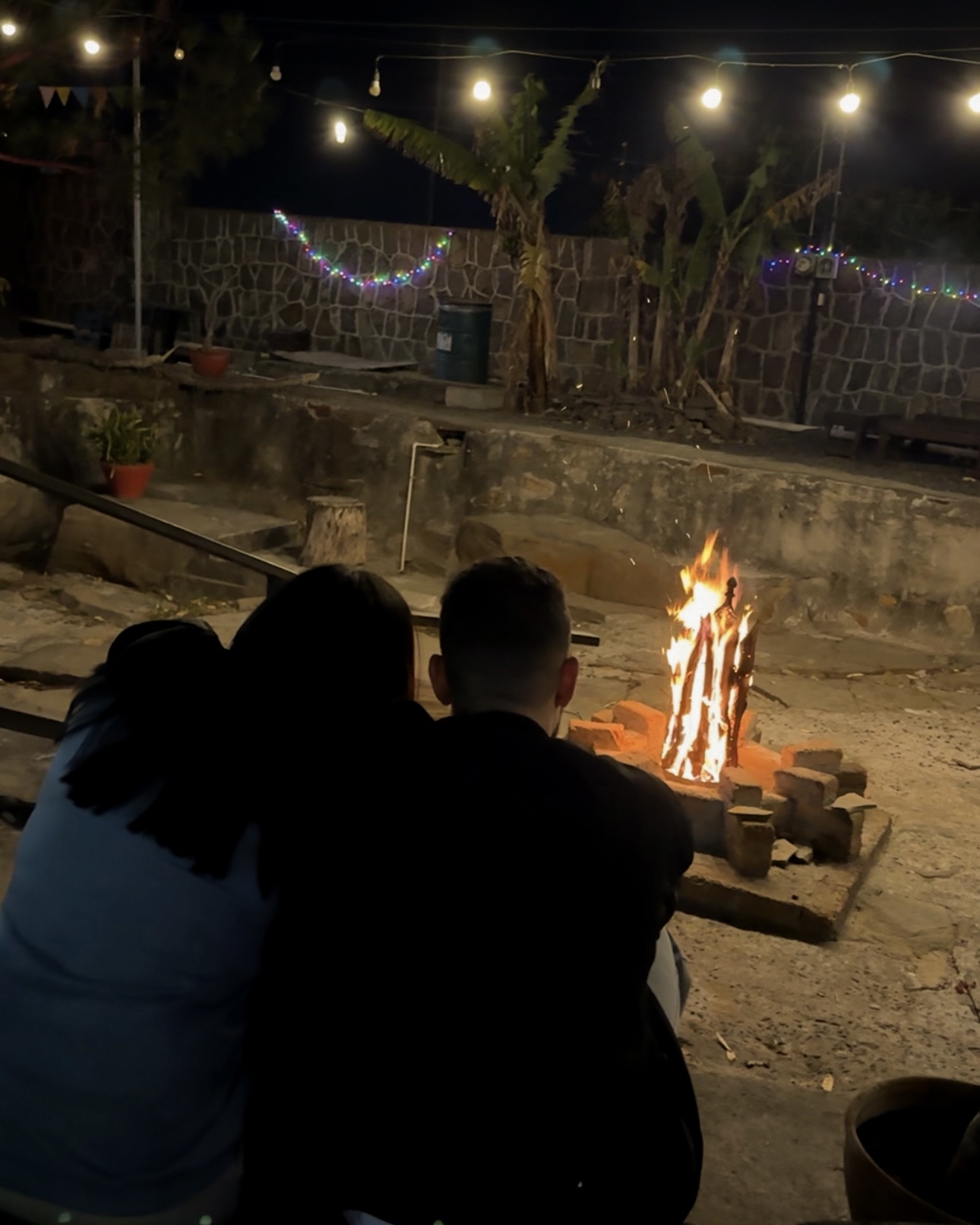 Pareja disfrutando de la fogata bajo luces de cadena en el patio del parque.